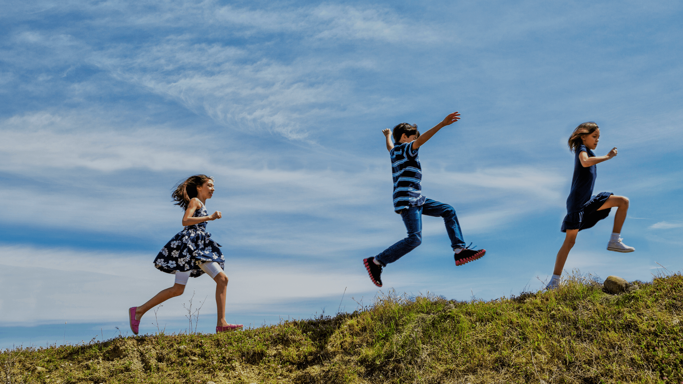 Three primary aged children running and jumping up a hill in the summer